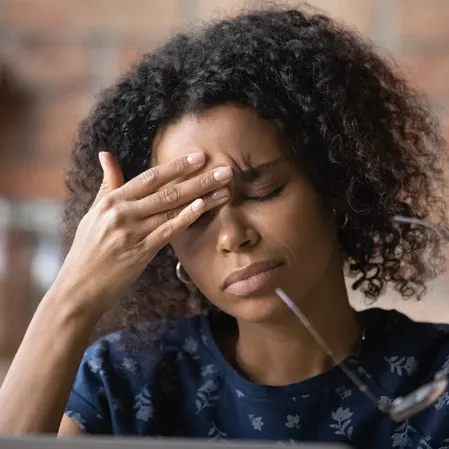 Person touching the forehead with one hand while holding eyeglasses, seated indoors with a blurred background.